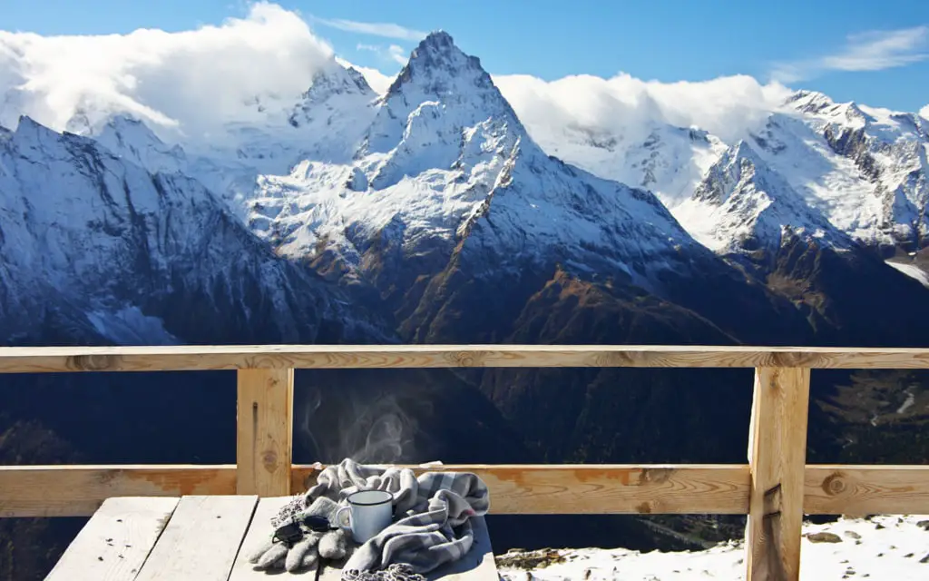 Hot tea with steam in tin mug, warm mittens, sunglasses and scarf on wooden table against snowy mountain peaks. Breakfast on terrace of cafe in mountain resort.